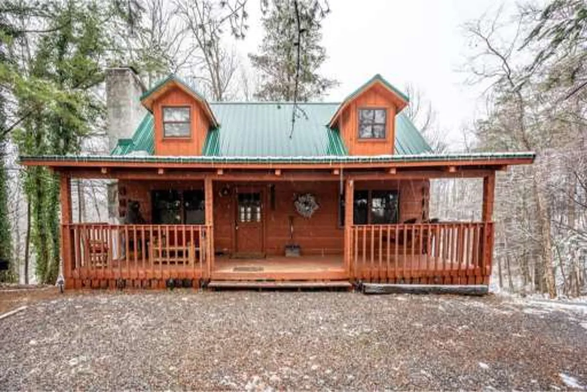 The hero image showcases the charming exterior of a Gatlinburg cabin, featuring a welcoming wooden porch, a striking green metal roof, a large front door adorned with a decorative wreath, and a picturesque snow-dusted setting that invites cozy gatherings. The porch hints at relaxing moments with hot cocoa while watching snowfall, and the spacious yard is perfect for family fun and building snowmen.