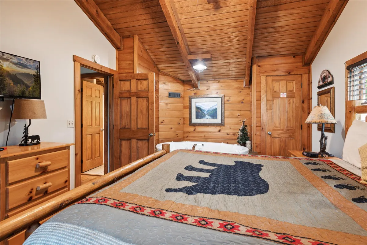 The scene features a king bedroom with a warm bear-themed quilt, sturdy wooden furniture, exposed ceiling beams, and tasteful wall art. The overall design exudes coziness and mountain charm, inviting guests to snuggle in and savor the tranquil surroundings.
