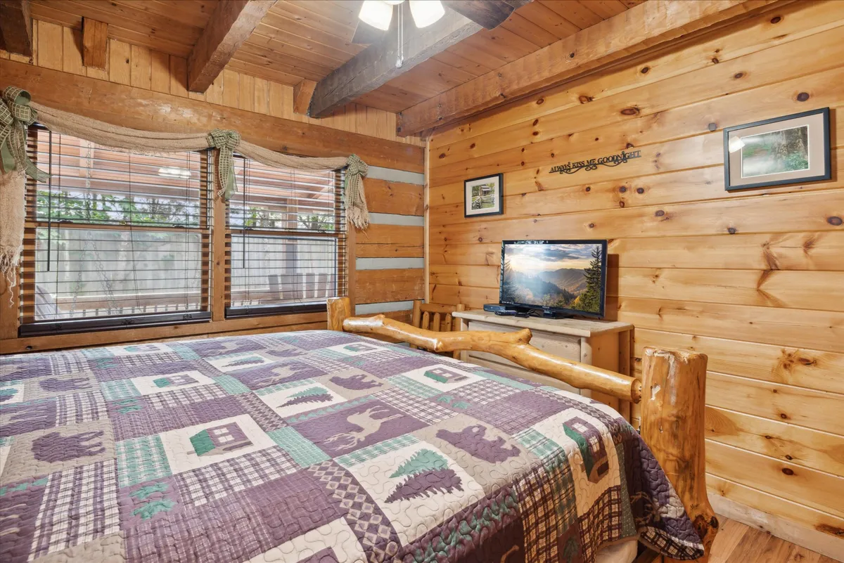 This image captures a king bedroom featuring a log bed adorned with a plush quilt, natural wood finishes, and large windows that allow plenty of natural light to filter in. The setting is warm and inviting, accentuated by the soft glow of sunlight and the comforting ambiance created by rustic details and a television for relaxation.