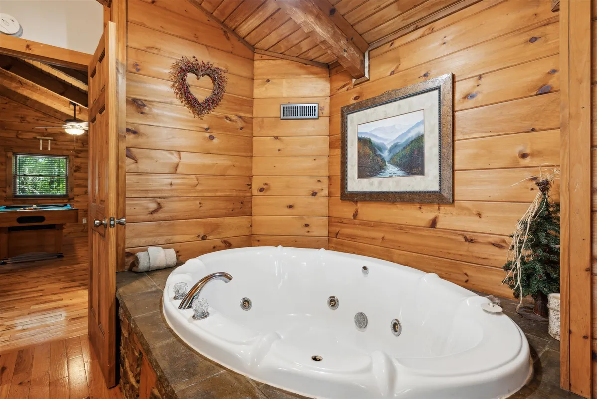 A tranquil bathroom scene featuring a whirlpool tub surrounded by warm wooden walls, with framed artwork and a decorative wreath adding a charming touch. The setting radiates serenity and romance, ideal for relaxing soaks in privacy.