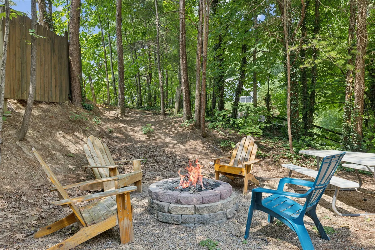 An inviting outdoor community fire pit encircled by wooden chairs and a picnic table, surrounded by tall trees. The setup is perfect for group gatherings, storytelling, and relaxing evenings under the stars, embodying warmth and camaraderie.