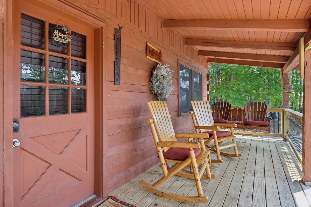 The cozy exterior of the cabin is shown, with prominent wooden rocking chairs on the deck and warm decorative touches near the front door. The deck invites families to relax, socialize, or watch kids play in the yard, surrounded by tall trees.