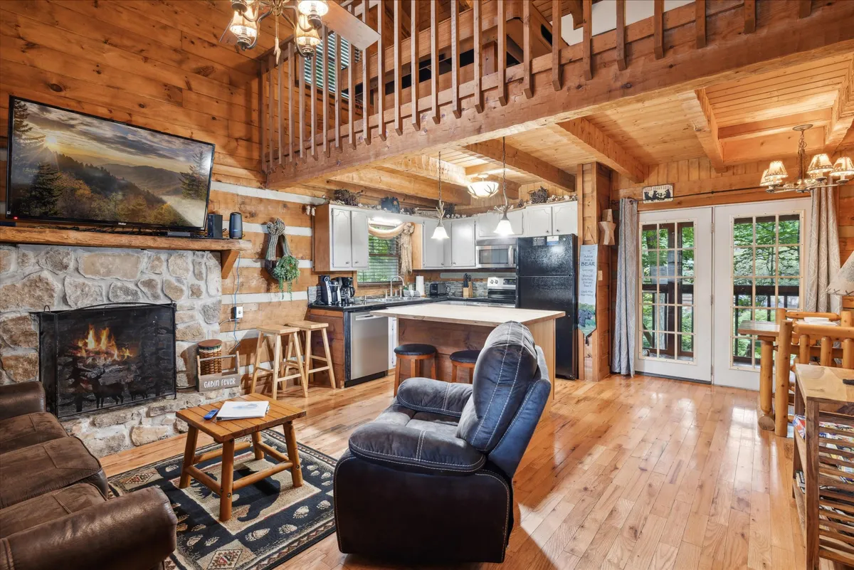 This image captures an open-concept living area with a cozy fireplace as a focal point, a comfortable sofa, a kitchen island, large windows that invite natural light, and a glimpse of the loft space above. The warm wood tones and rustic finishes set a welcoming and charming atmosphere.