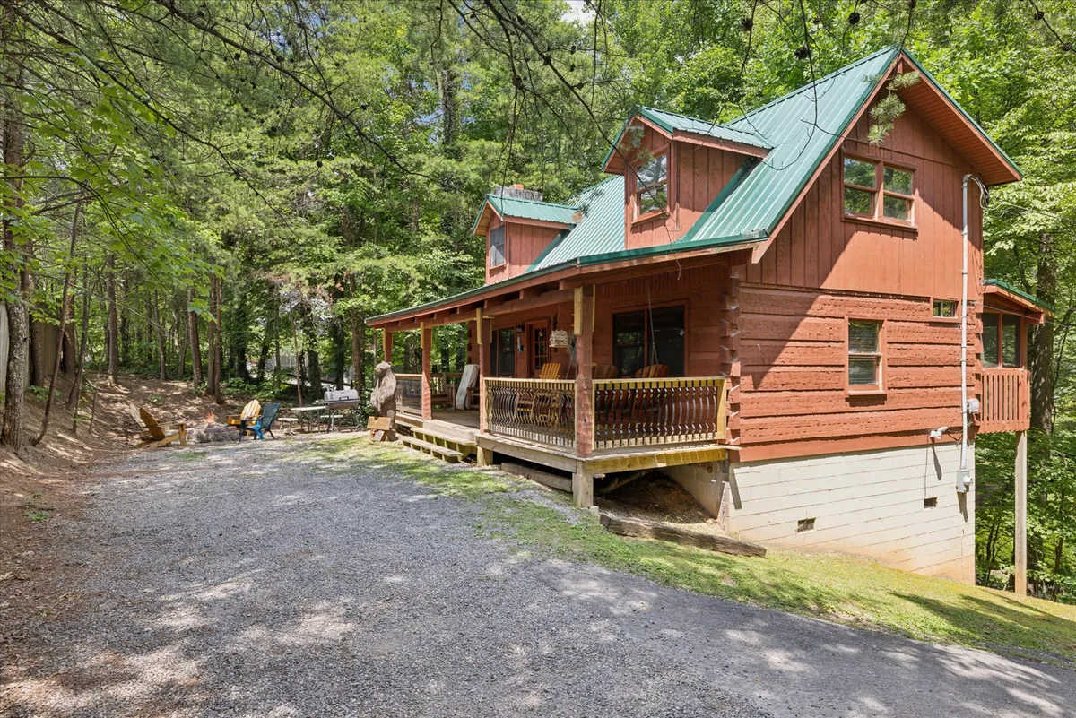 A rustic wooden cabin with a green metal roof sits nestled among tall trees. The wraparound porch features inviting spaces for sitting and enjoying the surrounding natural scenery. The peaceful, secluded setting is complemented by a yard ideal for gathering and a visible fire pit area, enhancing the romantic and private atmosphere.