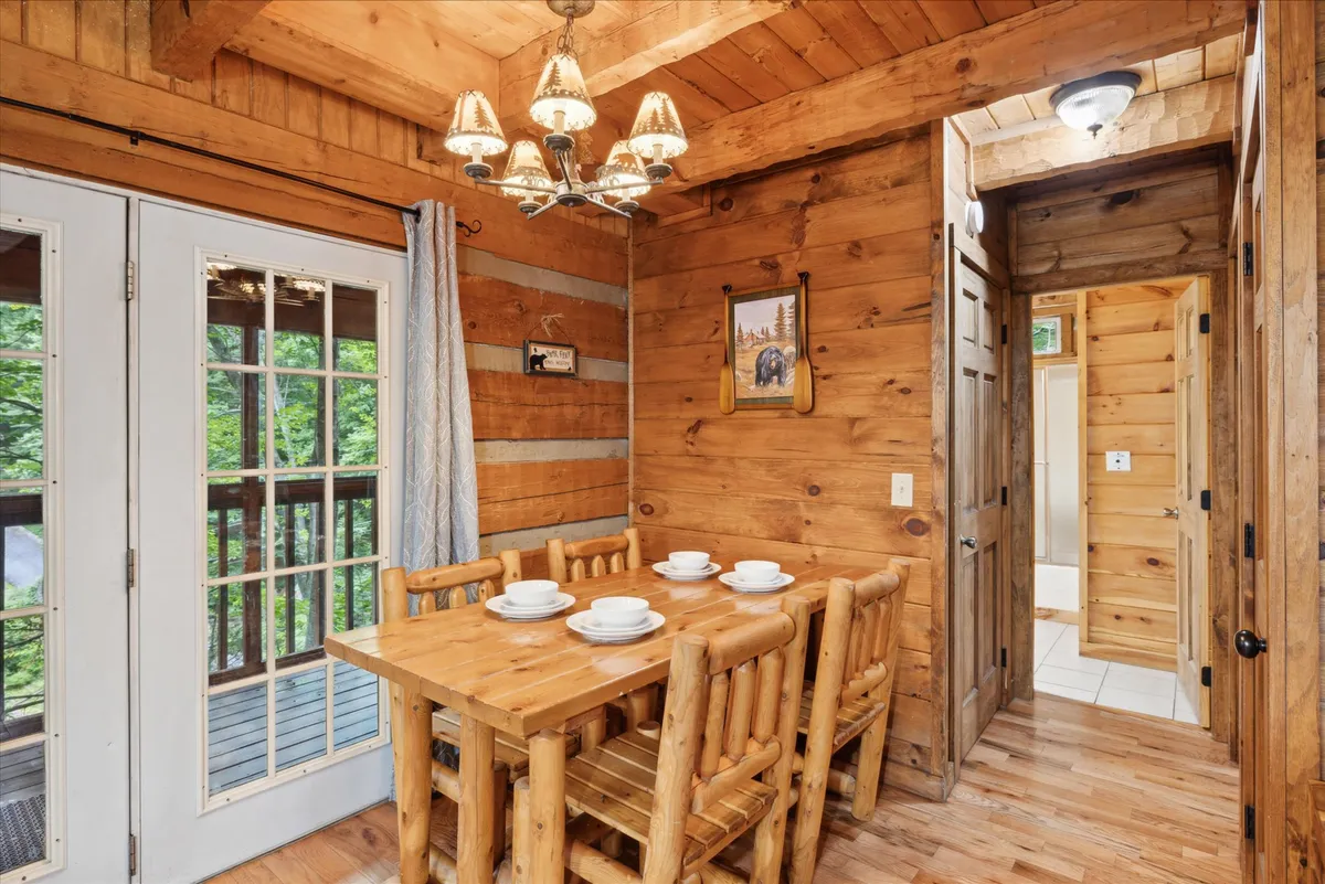 This image highlights a rustic wooden dining table set beneath a charming chandelier, flanked by large windows that bathe the room in natural light. The wood-paneled walls and intimate setting make it ideal for family dinners or relaxed gatherings.