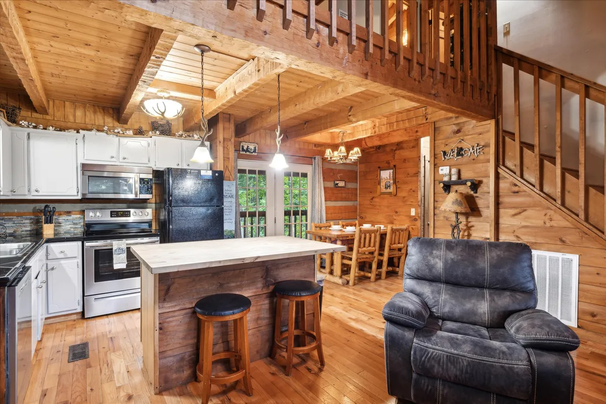 The photo focuses on a warm and functional kitchen area, featuring a kitchen island, black appliances, a wooden dining table, and a cozy recliner chair. The layout fosters connection between cooking, dining, and relaxing, all within a homey wood-paneled backdrop.